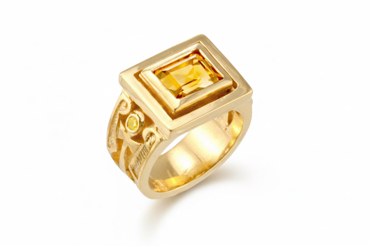 A men's gold signet ring featuring a large square-cut citrine stone, and ornate detailing that means "living in truth" on the band.