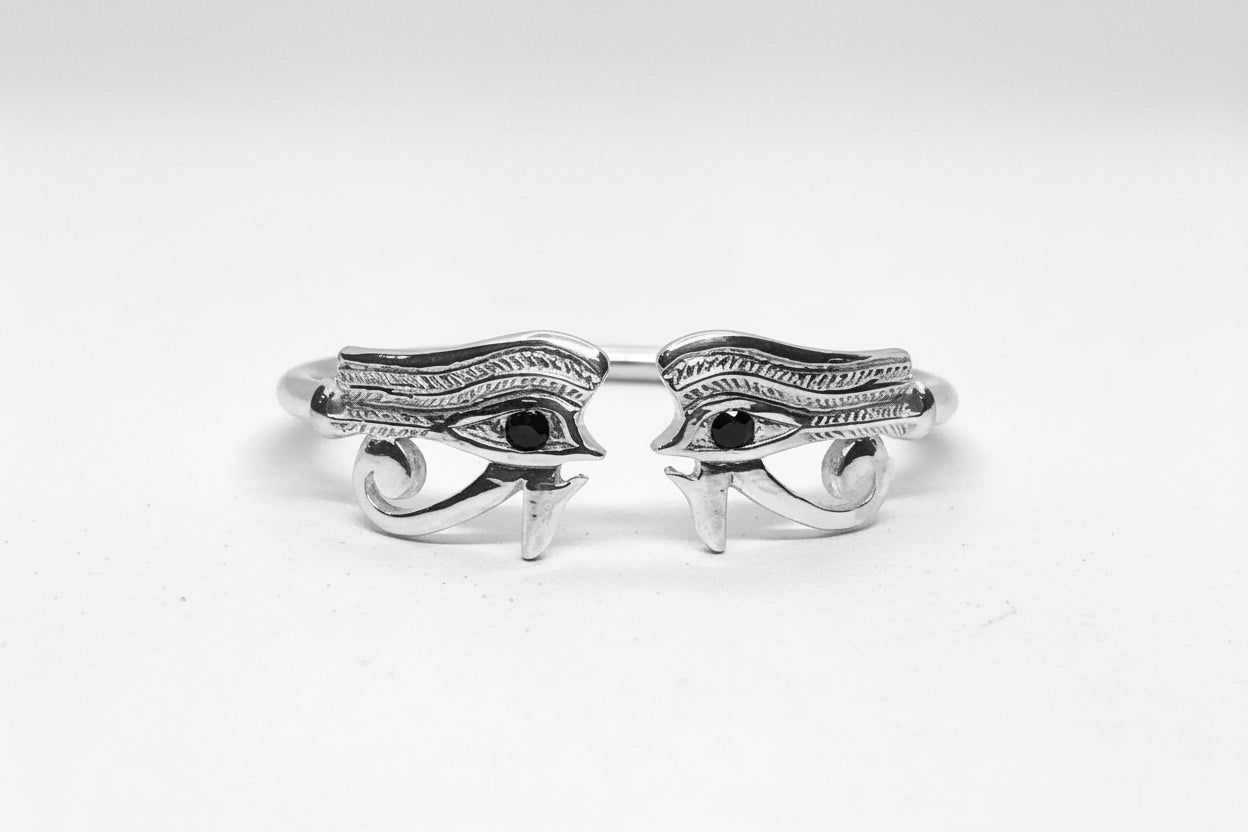 A sterling silver bangle featuring a hand-carved design resembling the left and right eye of Heru (Horus) with onyx stones in the eyes on a white background.
