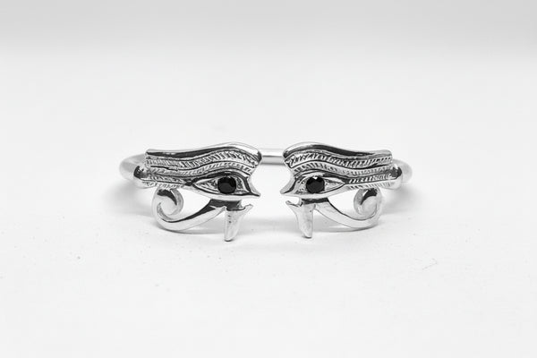 A sterling silver bangle featuring a hand-carved design resembling the left and right eye of Heru (Horus) with onyx stones in the eyes on a white background.