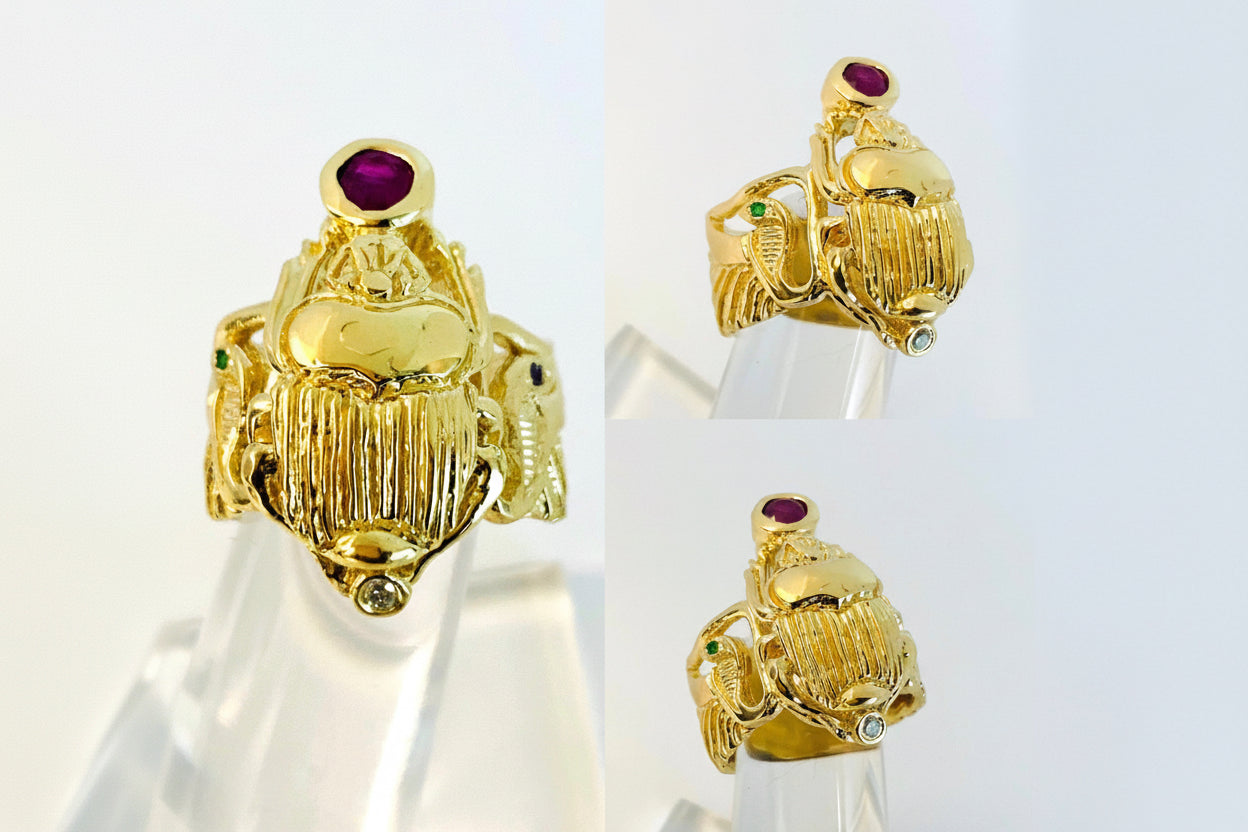 A 14k yellow gold ring with a scarab design, featuring a ruby and diamond, displayed on a clear stand.